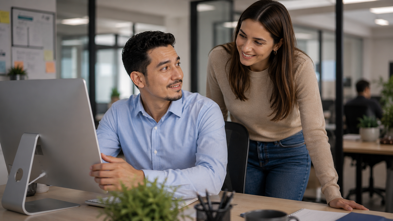 Homem sentado diante do computador mostra a tela para uma colega de trabalho em um escritório moderno, enquanto ela revisa um e-mail profissional em espanhol com um sorriso discreto.